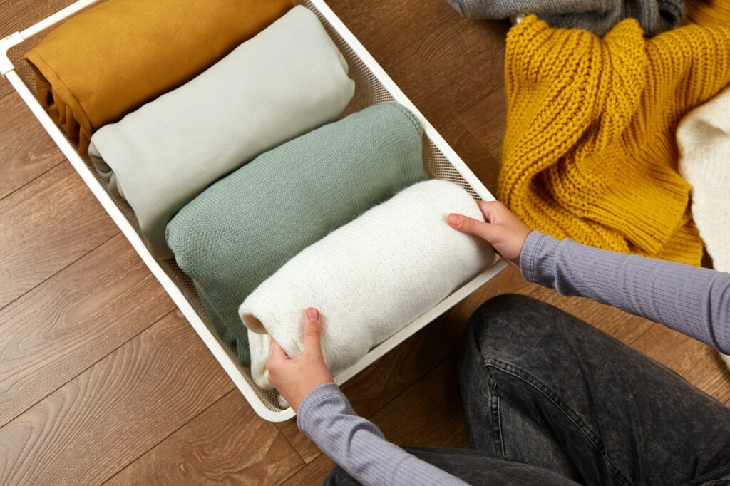 The hands of a woman neatly folding knitted clothes into a metal laundry basket